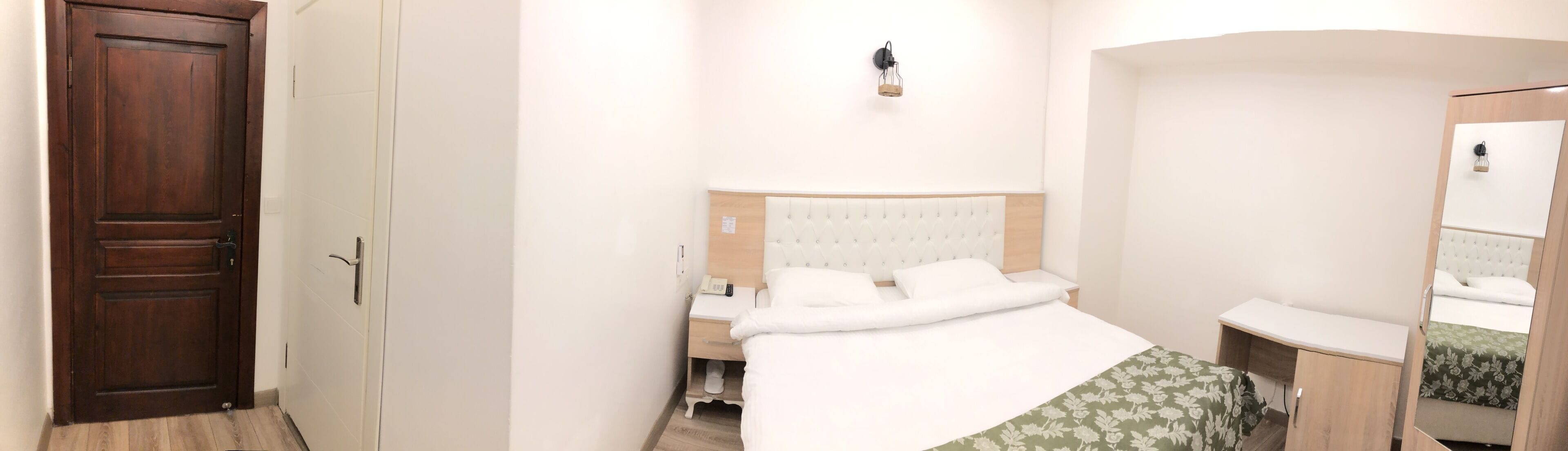 A hotel room with a wooden door, a bed with white bedding, a bedside table with a lamp, and a small wooden cabinet.