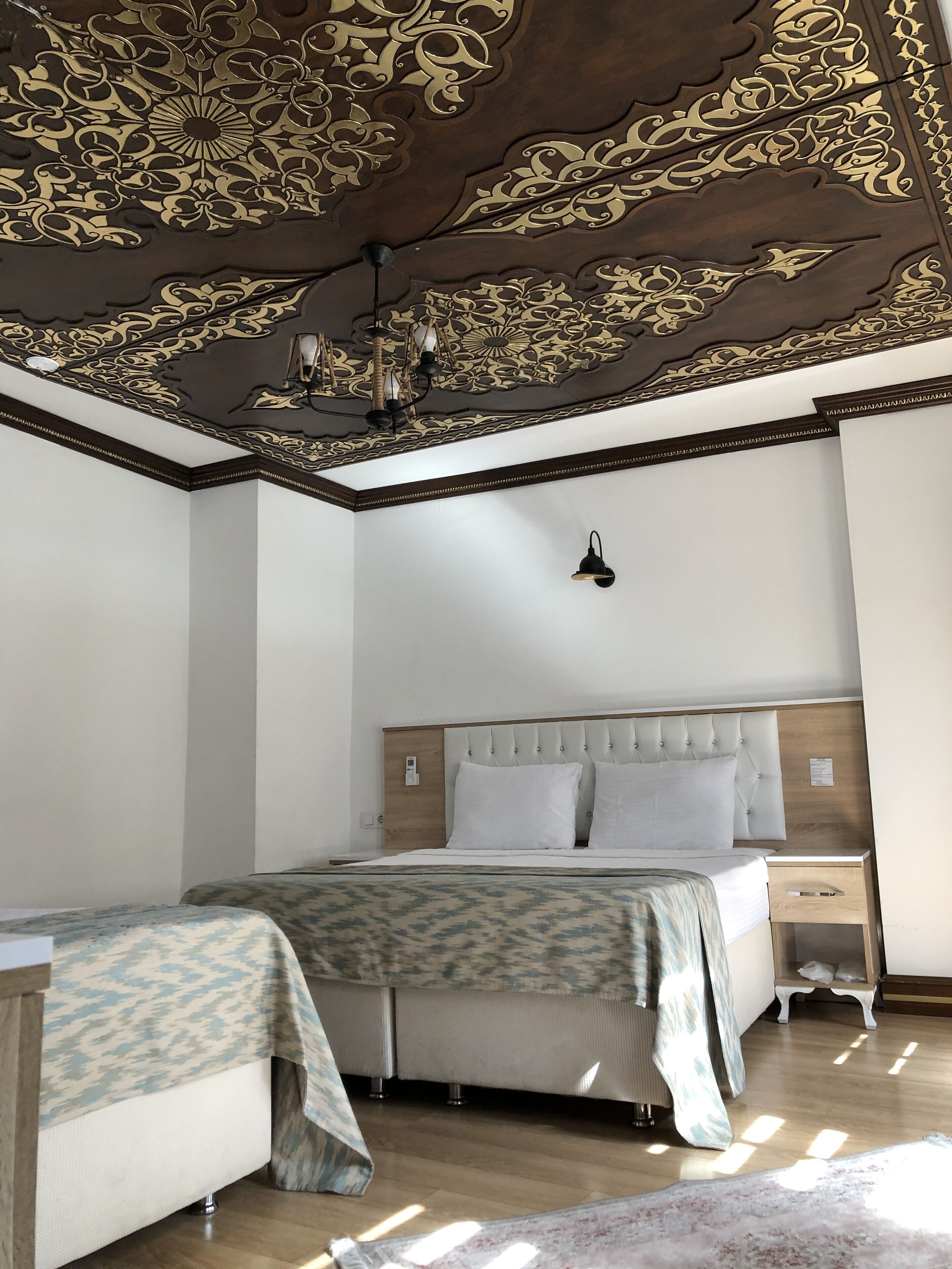 Ein Schlafzimmer mit einem großen Bett, Nachttischen, einem Beistelltisch, einer gemusterten Bettdecke und einer Decke mit aufwendigen Verzierungen.
