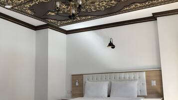 Ein Schlafzimmer mit einem großen Bett, Nachttischen, einem Beistelltisch, einer gemusterten Bettdecke und einer Decke mit aufwendigen Verzierungen.