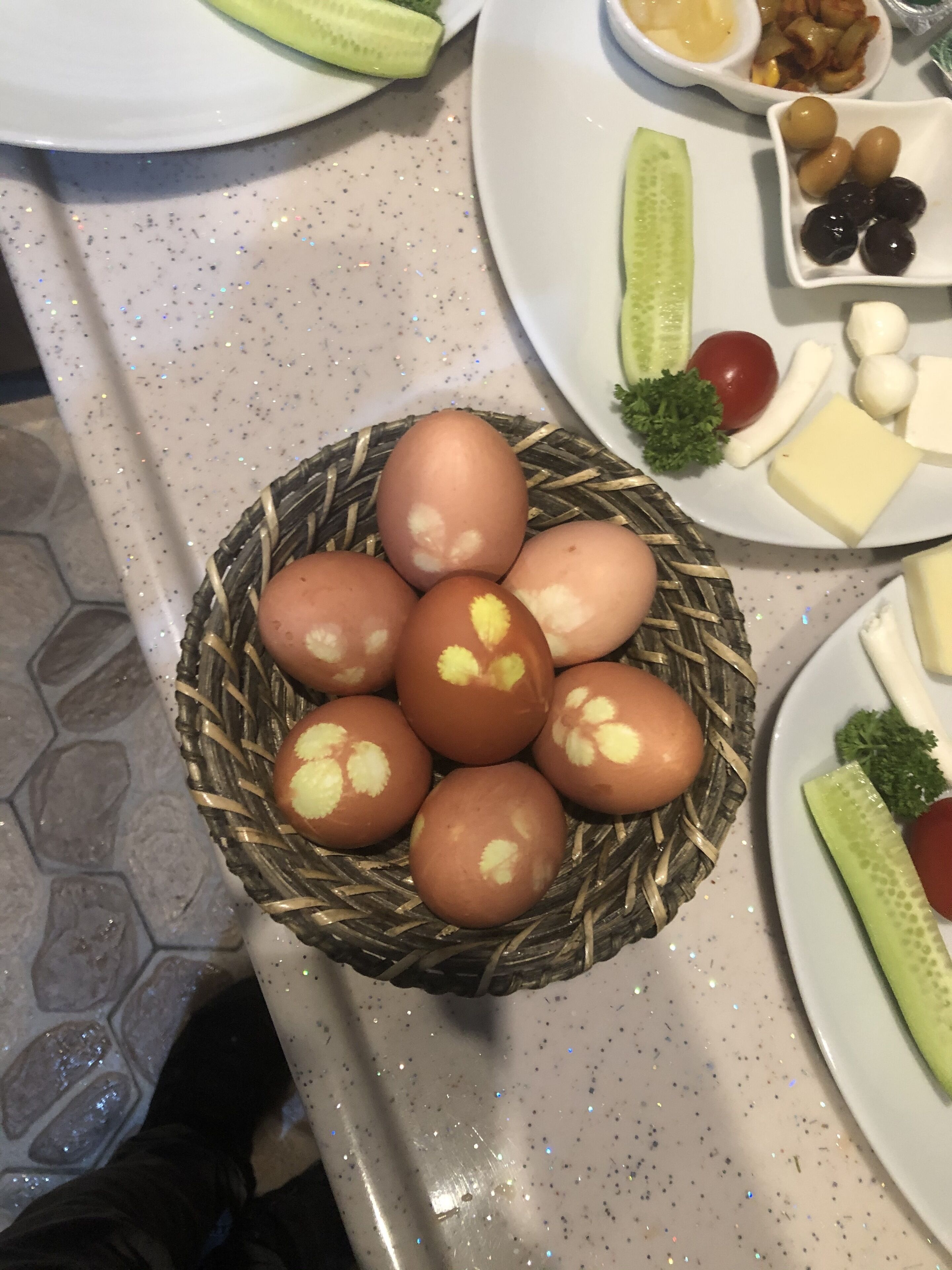 A table with a basket of decorated eggs, a plate with cheese, olives, and vegetables, and a plate with a cucumber and tomato.