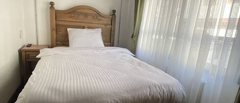 Un dormitorio con cabecera de madera, cubrecama blanco, mesita de noche con control remoto y ventana con cortinas traslúcidas.