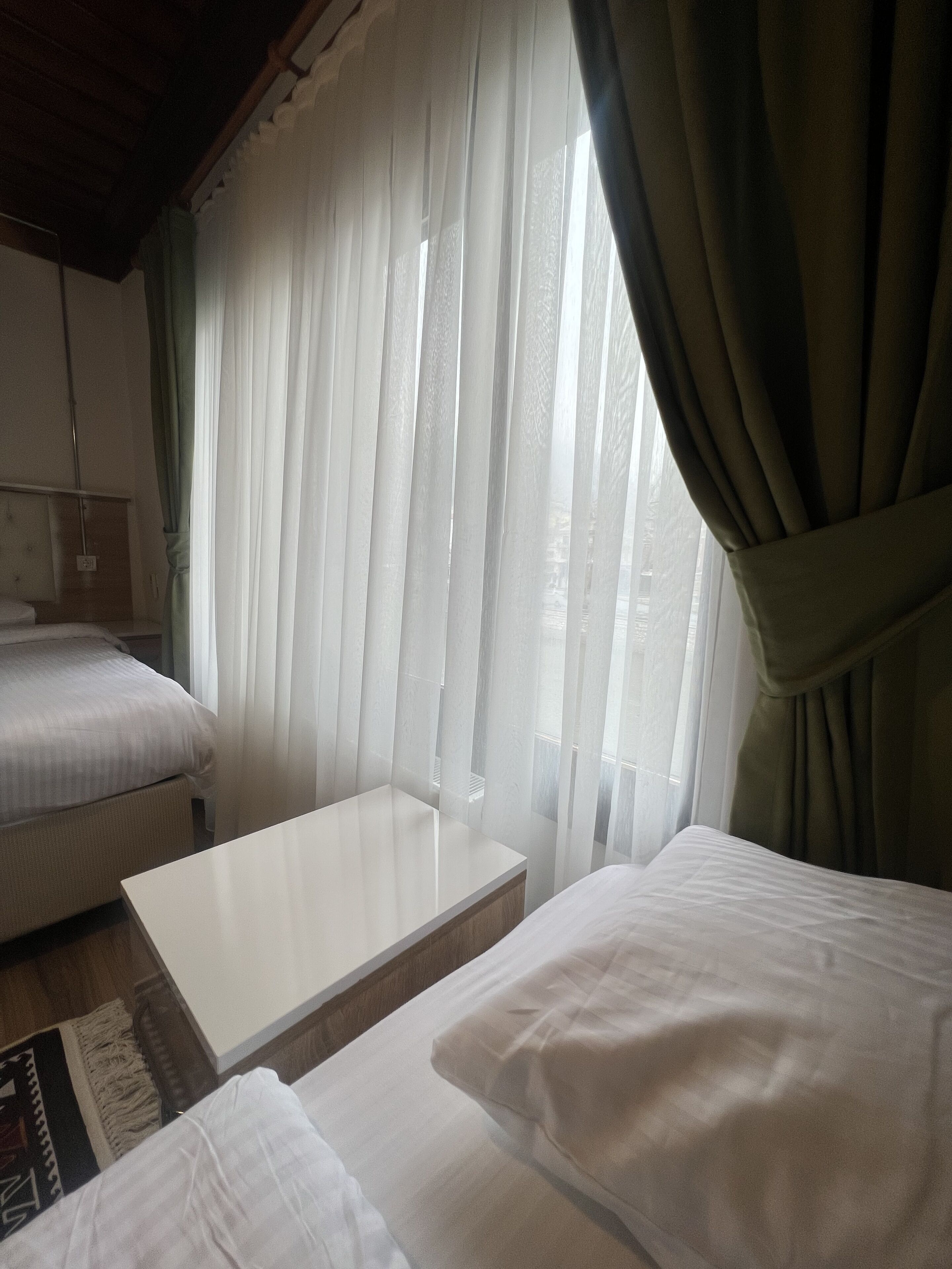 A hotel room with two beds, a small table, and sheer curtains.
