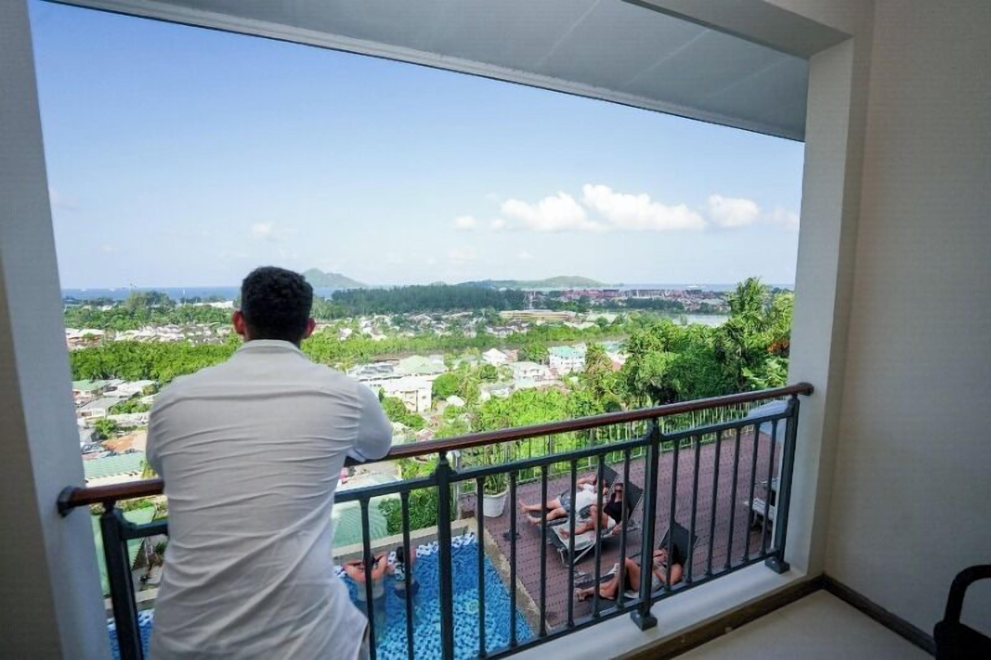 Een man staat op een balkon met uitzicht op een stadsgezicht met gebouwen en groen.
