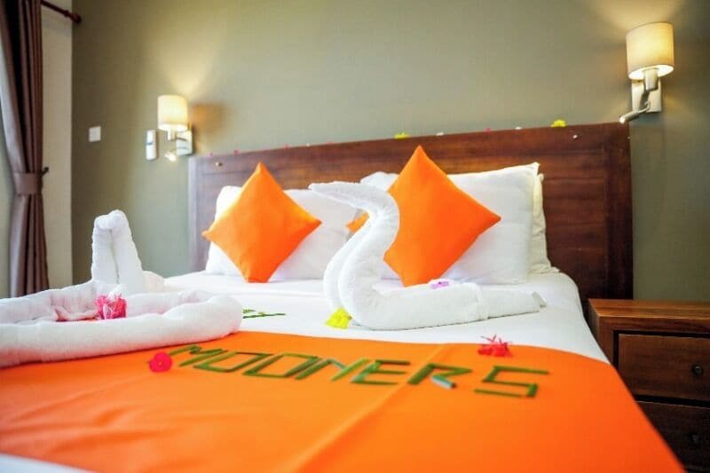 Una habitación de hotel con una cama decorada con un arreglo de toallas con forma de cisne, dos almohadas anaranjadas y un edredón con el texto 'MOONERS'.