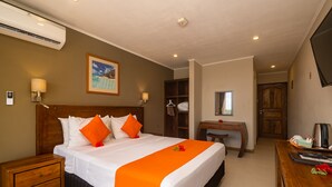 A hotel room with a large bed, a wooden nightstand, a desk, a chair, a TV, and a painting on the wall. - The Ridge Residence (Mahé Island)