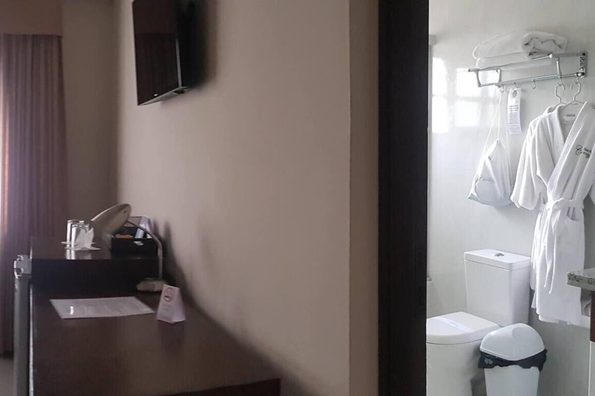 Habitación de hotel con cama, escritorio, silla, mesita, espejo, ventana con cortinas y baño con lavamanos e inodoro.