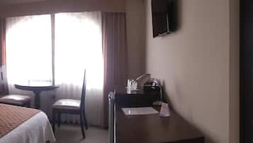 Une chambre d’hôtel comprenant un lit, un bureau, une chaise, une petite table, un miroir, une fenêtre avec des rideaux et une salle de bain avec un lavabo et des toilettes.