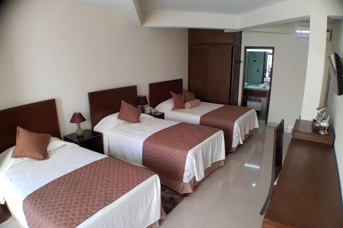 Habitación de hotel con tres camas, cada una con cabecero marrón y ropa de cama blanca. Hay un escritorio con lámpara y espejo.