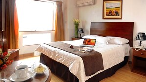 Habitación de hotel con cama, una laptop sobre la cama, una mesita de noche con lámpara, una pequeña mesa con una taza de café y un plato de comida, y una ventana con cortinas.