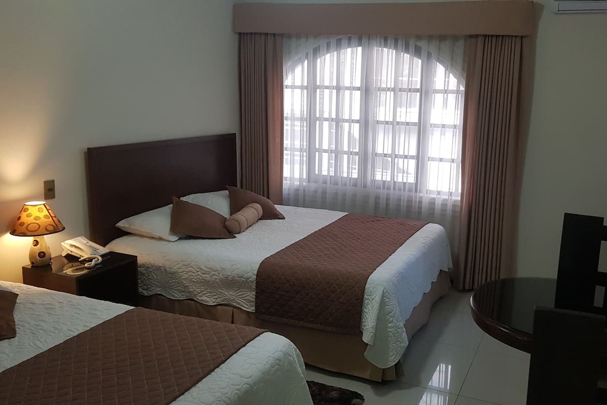 Una habitación de hotel con dos camas, una mesita de noche con lámpara y teléfono, una ventana con cortinas y una pequeña mesa con una silla.