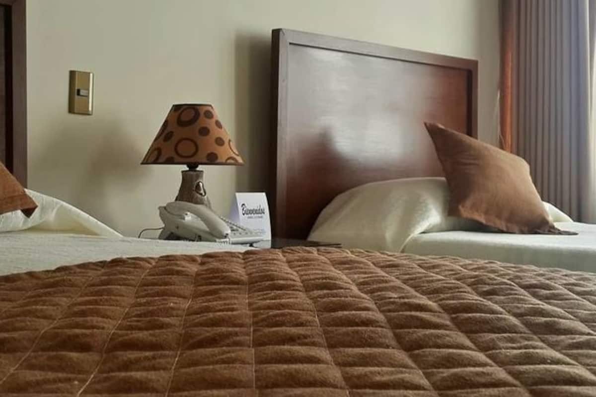 Habitación de hotel con dos camas, un edredón marrón acolchado, una lámpara con pantalla de lunares y un cartel en la mesita de noche.
