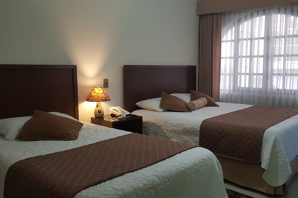 Habitación de hotel con dos camas, una mesita de noche con lámpara, teléfono y un ventanal con cortinas.