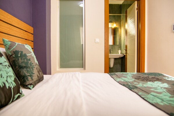 Une chambre à coucher avec un lit, des oreillers et une porte-fenêtre coulissante donnant sur une salle de bain équipée d’un lavabo et d’un miroir.