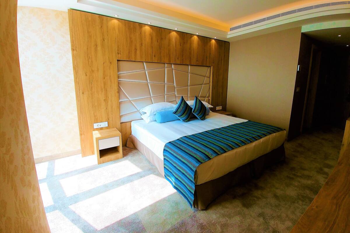 A modern hotel room with a large bed, a wooden headboard, a small bedside table, and a carpeted floor.