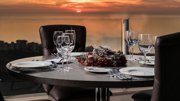 A dining table set for four with wine glasses, plates, and cutlery, overlooking a cityscape at sunset. - Maximus Hotel Byblos (Blat)