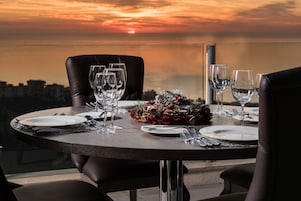 Mesa de comedor para cuatro personas, con copas de vino, platos y cubiertos, con vistas a un paisaje urbano al atardecer.
