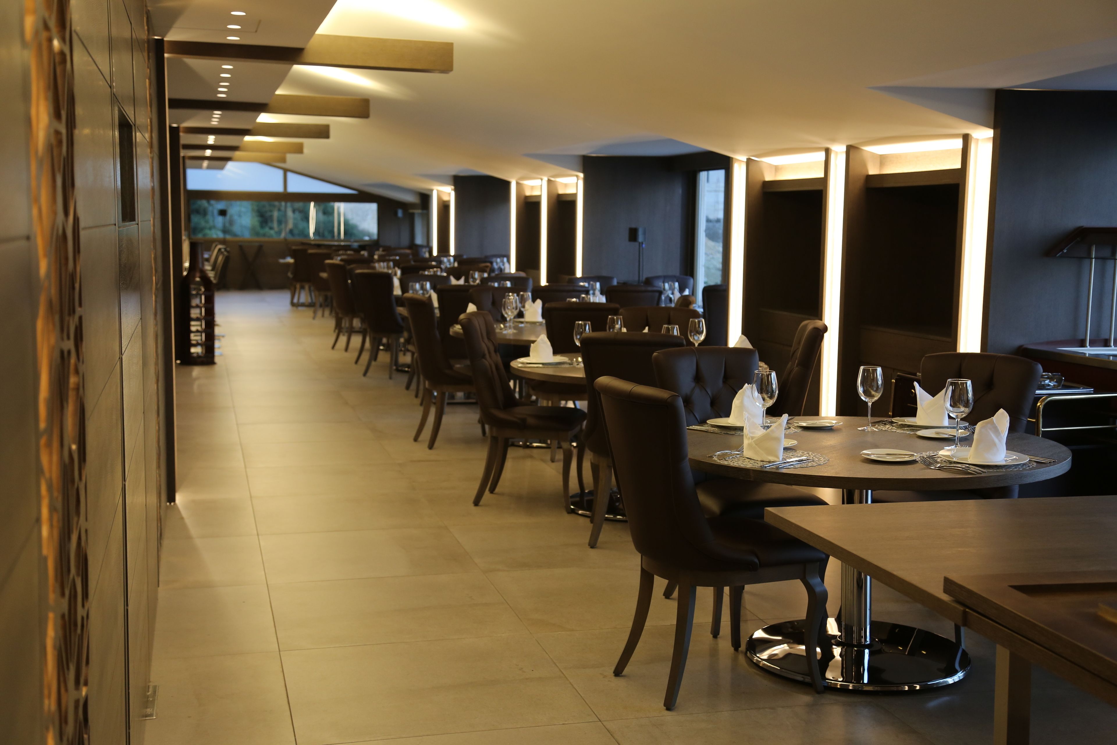 Un restaurante moderno con mesas y sillas largas, un suelo de baldosas limpias y ventanas grandes que dejan entrar luz natural.
