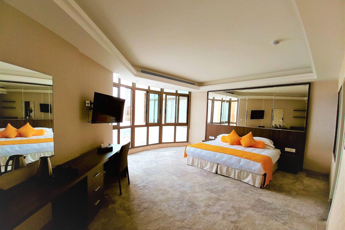 A hotel room with two beds, a desk, a chair, a television, and large windows.
