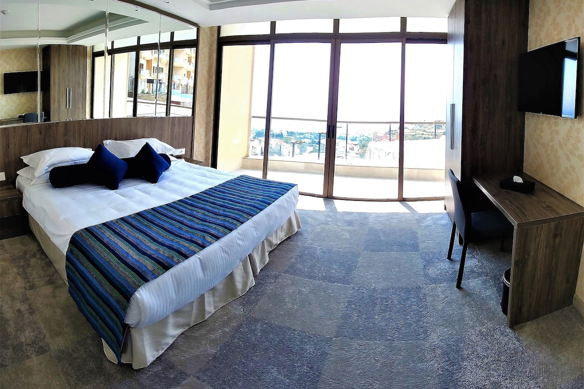 A hotel room with a large bed, a desk, a flat-screen TV, and a balcony with a city view.