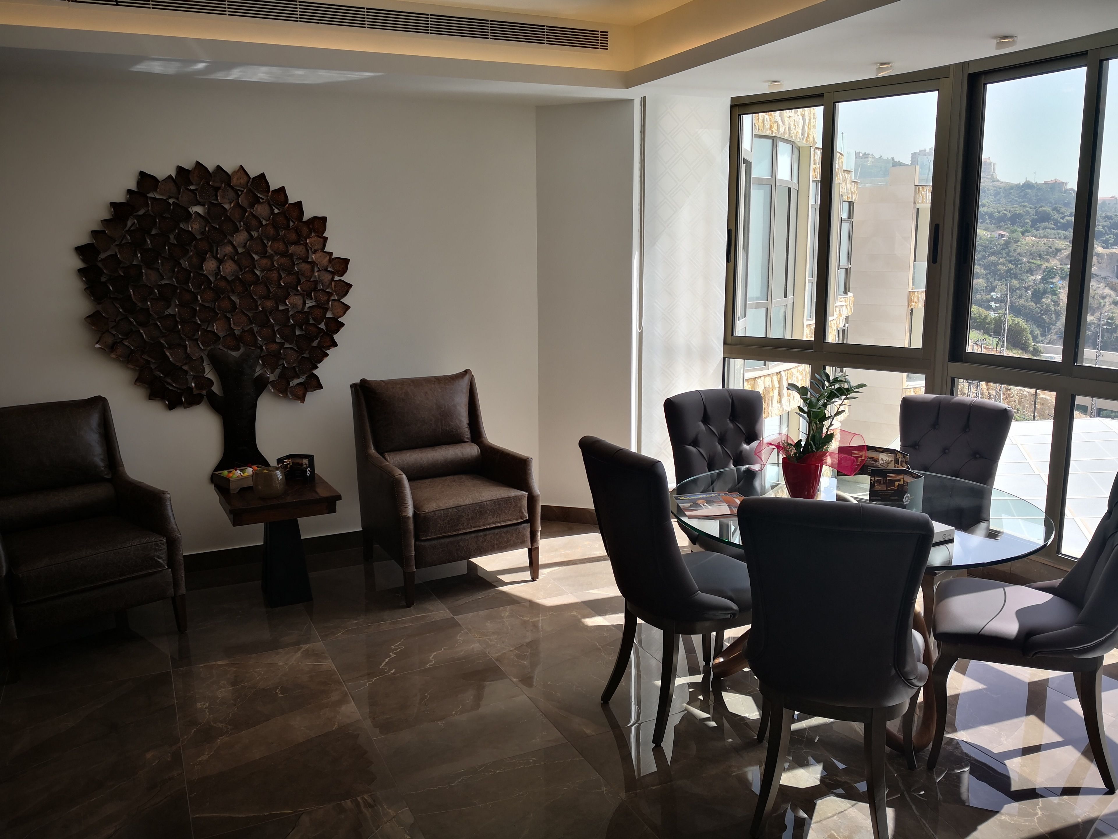 A modern room with a glass table surrounded by chairs, a decorative tree wall art, and a view of buildings through large windows.