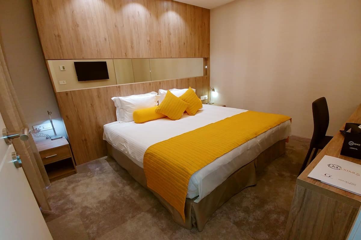 A hotel room with a wooden headboard, a bed with white bedding and a yellow throw, a bedside table with a lamp, and a desk with a chair.