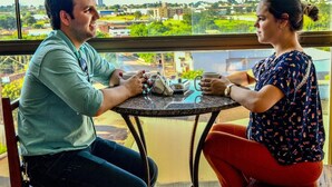 Two people seated at a round table with a view of a cityscape through large windows. - Hotel Califórnia (Jatai)