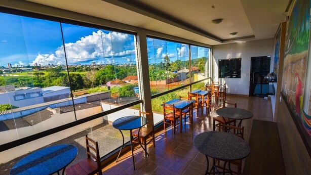 A spacious room with large windows offering a view of a cityscape and greenery. There are several wooden tables and chairs arranged for dining or sitting. - Hotel Califórnia (Jatai)