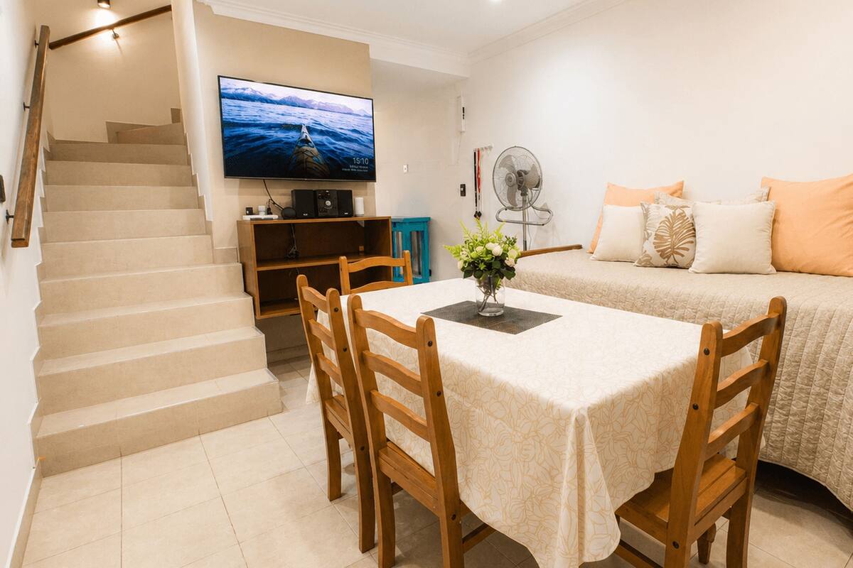 A hotel room with a dining table, chairs, a sofa, a TV, a fan, and a staircase.