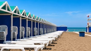 A beach with rows of white lounge chairs and blue umbrellas, a lifebuoy, and a lifeguard tower. - Casa Colinas (Mar del Plata)
