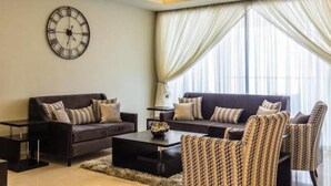 A modern living room with a dark sofa set, a coffee table, and patterned armchairs. A large wall clock is mounted on the wall. - Amara Suites - Eko Atlantic (Lagos)