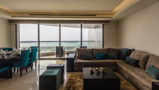 A modern living room with a large window overlooking the ocean, a dining area with teal chairs, and a cozy seating arrangement with a patterned ottoman. - Amara Suites - Eko Atlantic (Lagos)