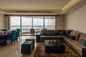 A modern living room with a large window overlooking the ocean, a dining area with teal chairs, and a cozy seating arrangement with a patterned ottoman. - Amara Suites - Eko Atlantic (Lagos)