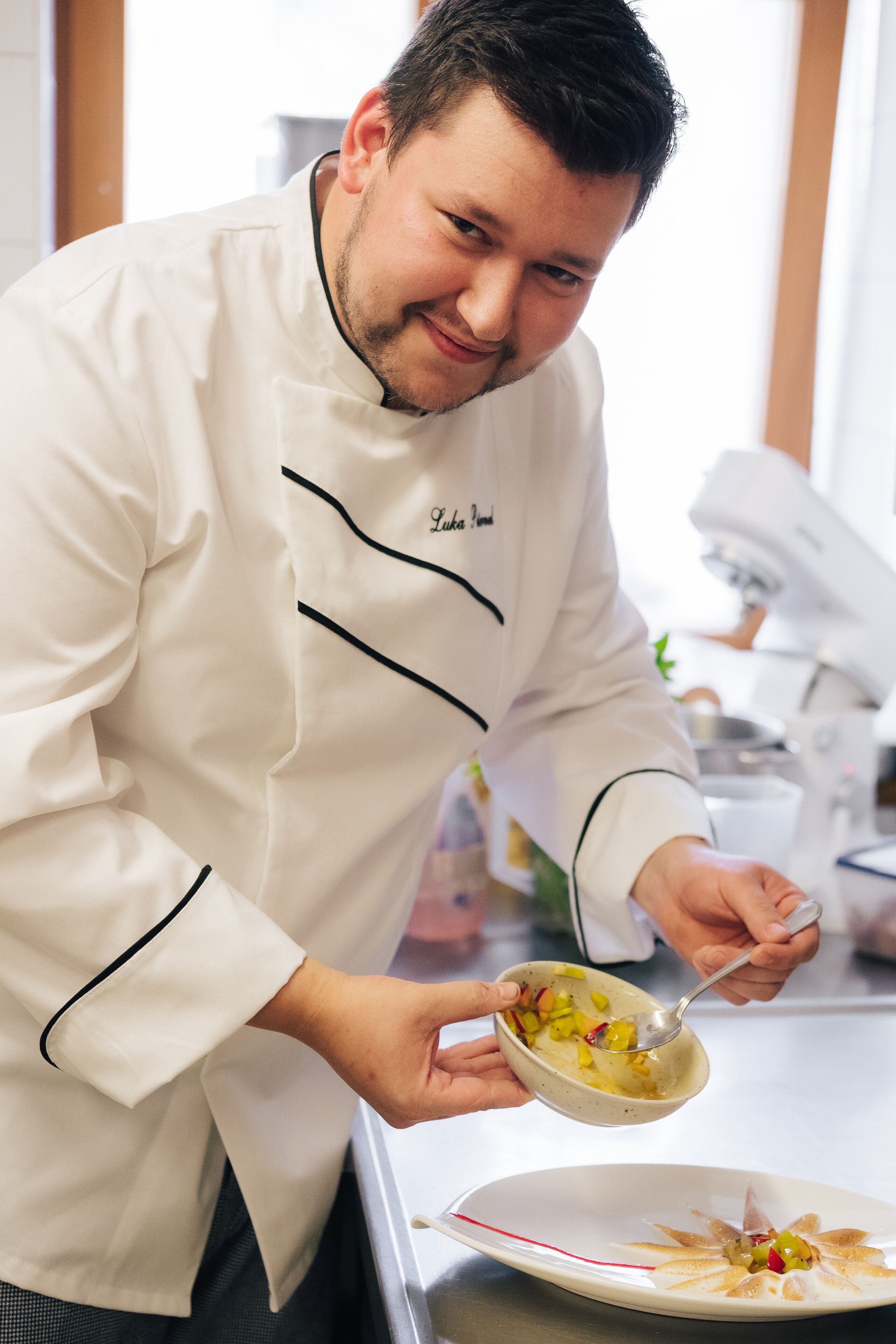A chef in a white uniform with black trim, holding a bowl of food and a spoon, standing in a kitchen with stainless steel appliances.