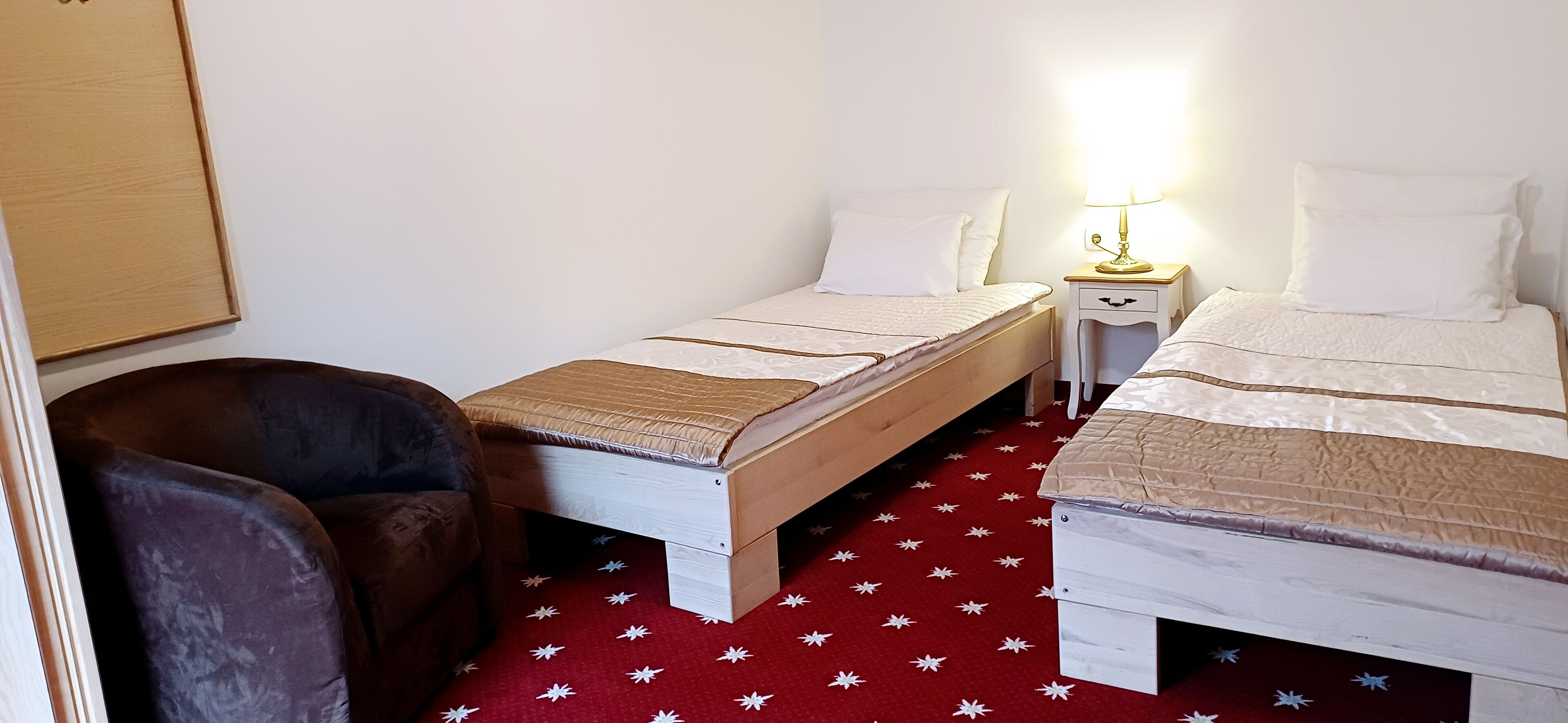 A small, cozy room with two single beds, a nightstand with a lamp, a chair, and a red star-patterned carpet.