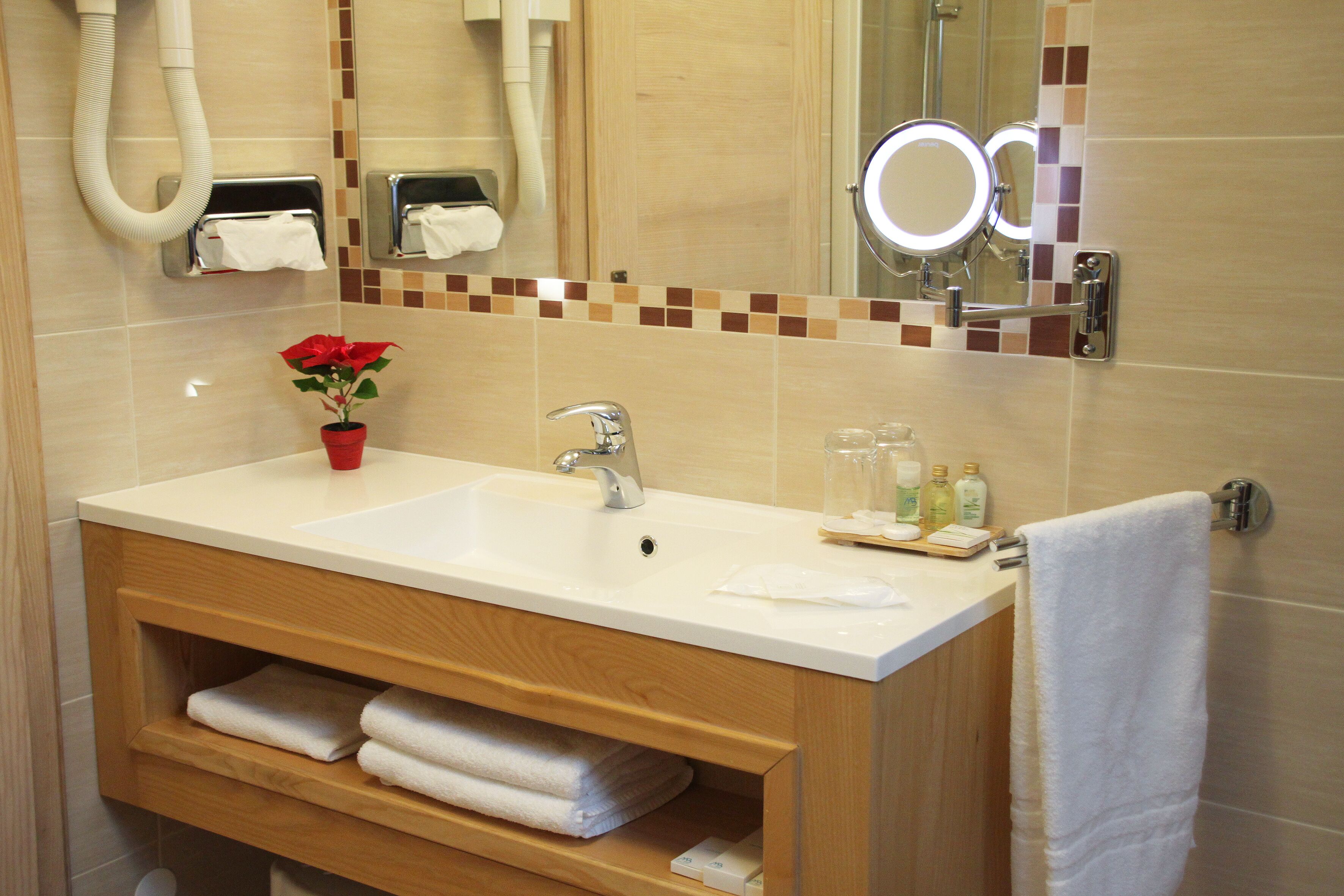 A bathroom with a white sink, wooden vanity, and a mirror with lights. There are towels and toiletries on the counter.