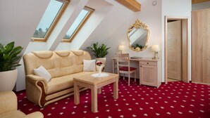 A cozy loft bedroom with a sloped ceiling, a beige sofa, a wooden desk, a chair, a mirror, and a red star-patterned carpet. - Hotel Planinka (Ljubno)