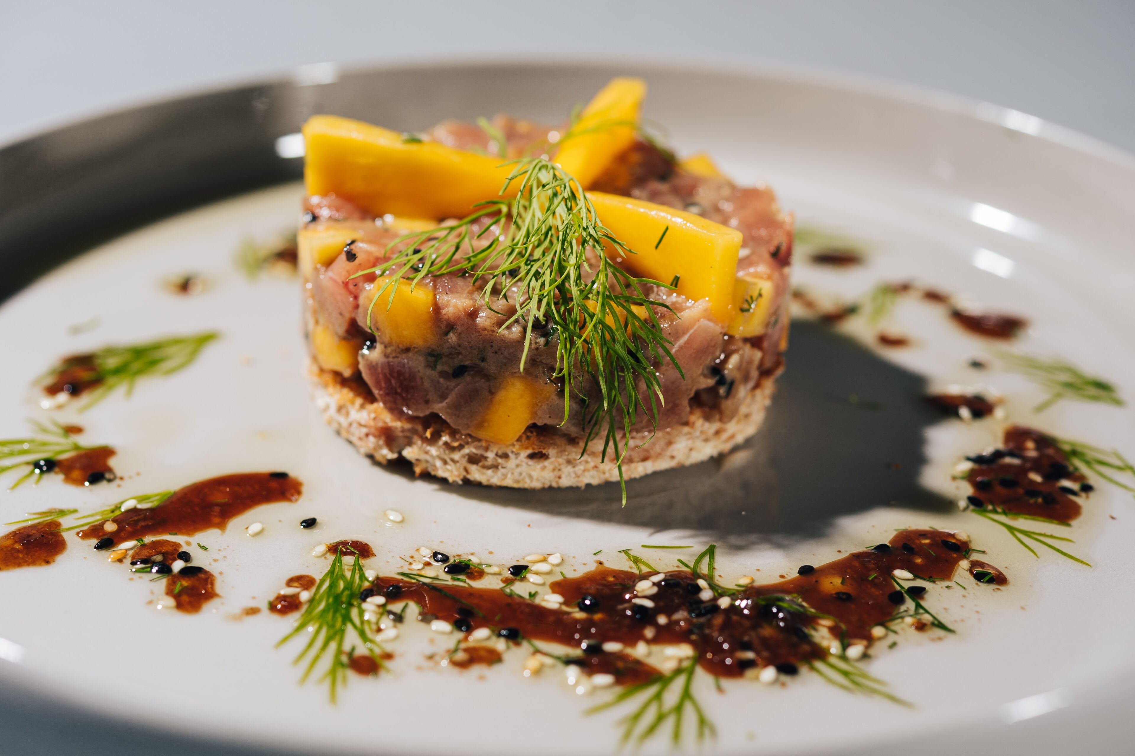 A dish with a slice of bread topped with meat and cheese, garnished with herbs, on a white plate with sauce and seeds around it.