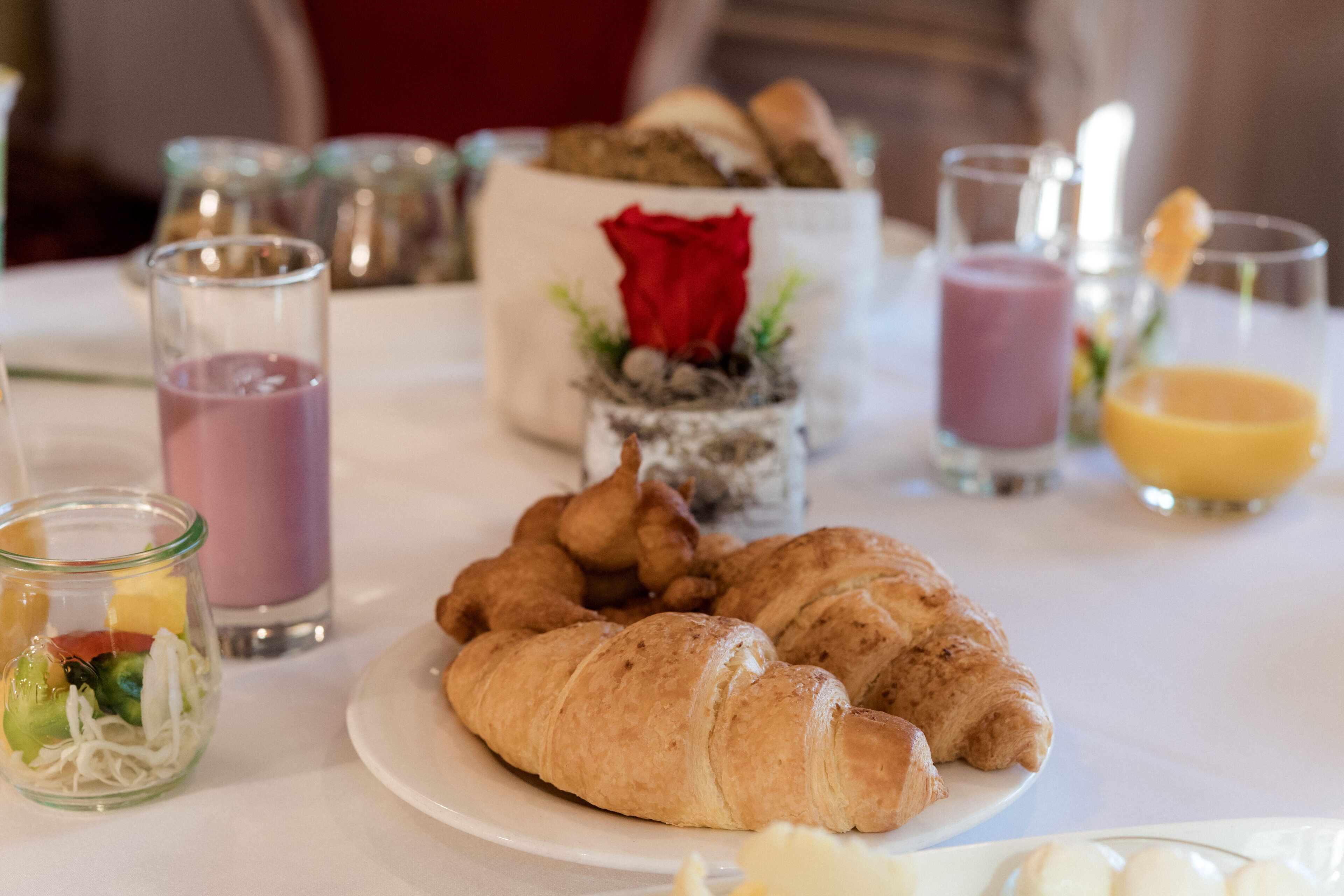 Aamiaispöytä, jossa on croissantteja, lasi mehua ja kynttilänjalka, jossa on ruusu.