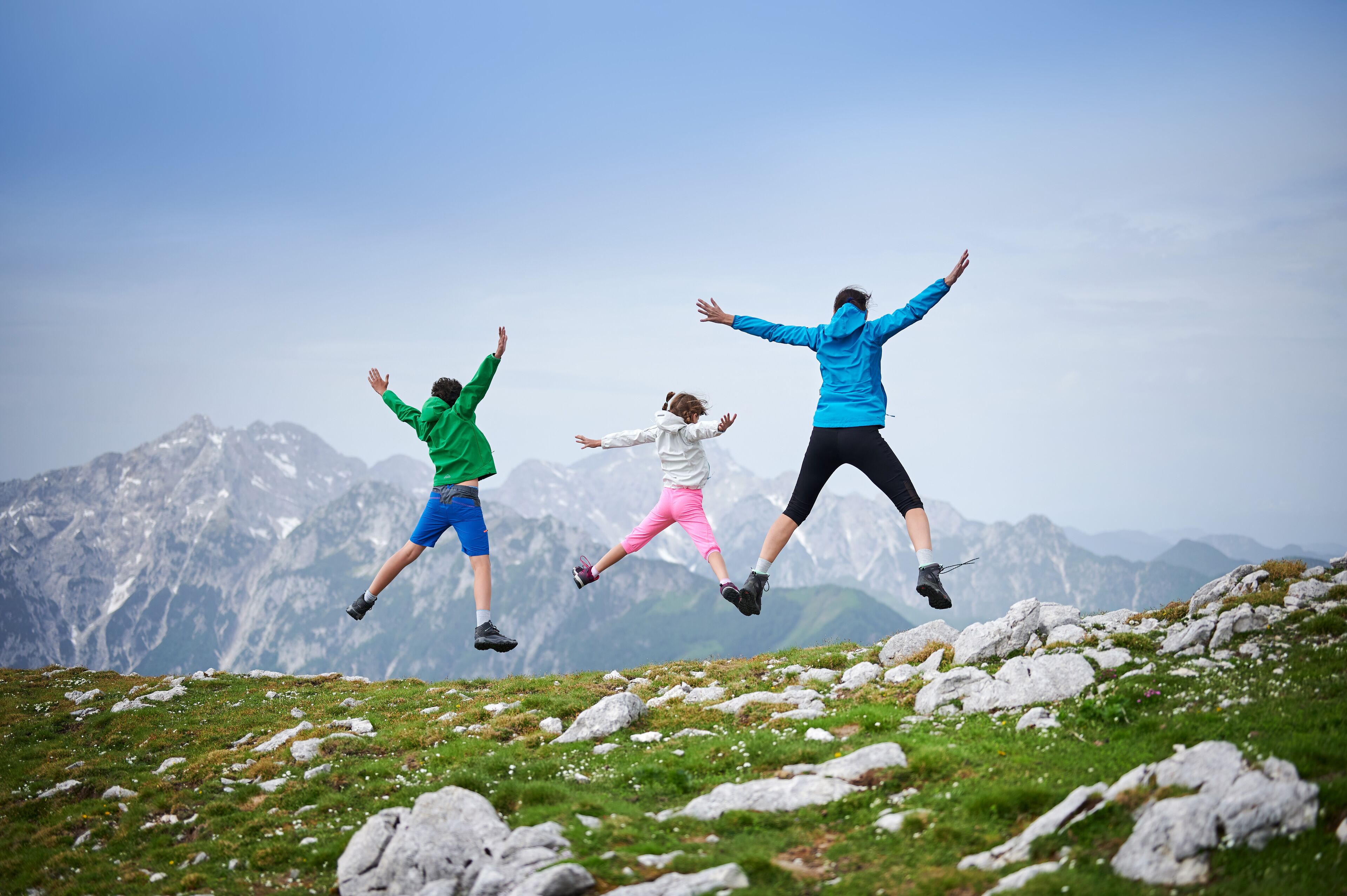 Three individuals jumping on a grassy mountain peak with a mountainous backdrop.