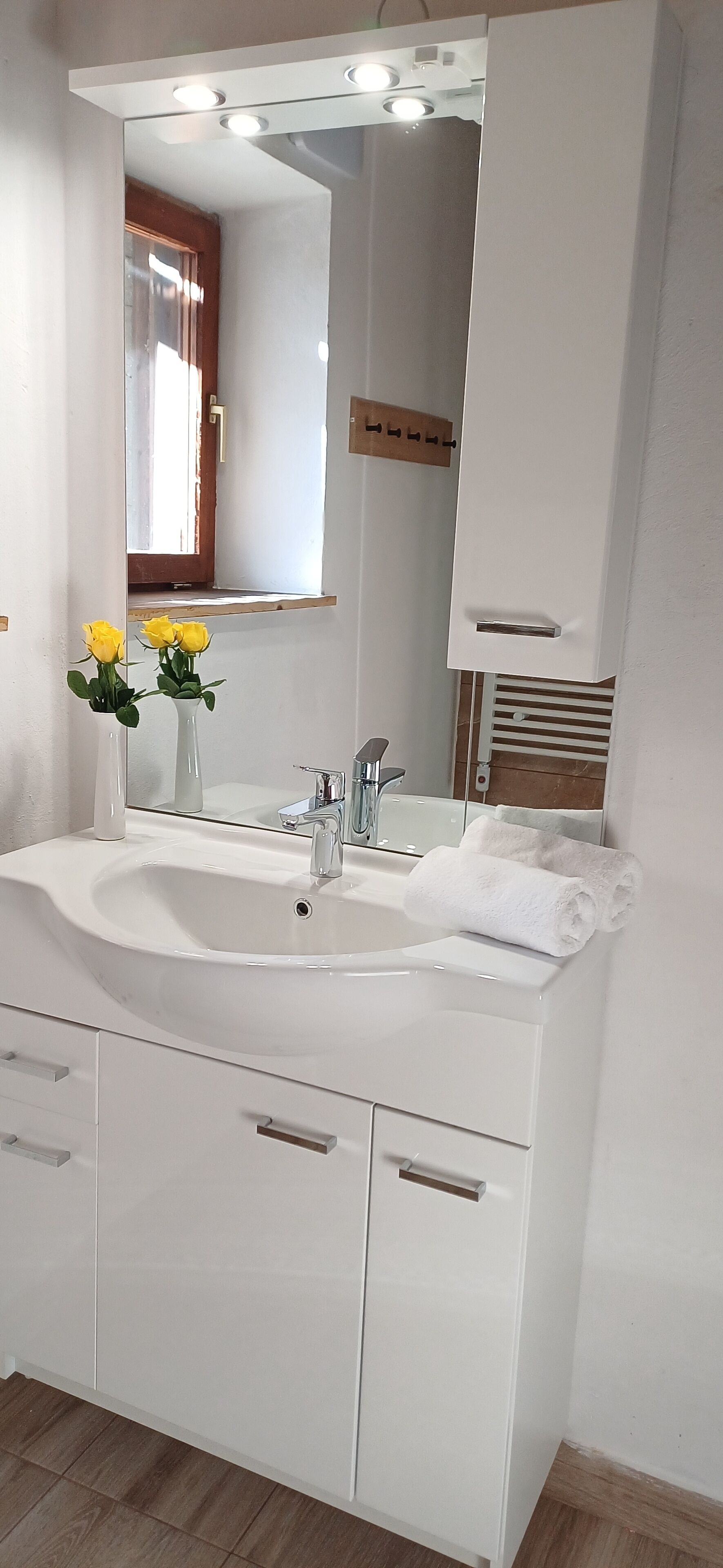 Une salle de bain avec un lavabo blanc, un miroir et une commode. Un vase contenant des fleurs jaunes est placé à gauche, et une serviette à droite.