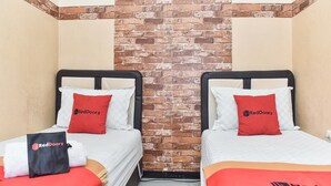 A hotel room with two beds, each with a white bedspread and a red pillow featuring the RedDoorz logo. The room has a brick accent wall and beige walls. - RedDoorz near Stasiun Kota Baru Malang (Malang)