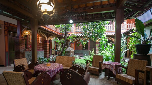 An indoor patio with wooden furniture and a tiled floor, surrounded by greenery and a view of a traditional building. - RedDoorz near Stasiun Kota Baru Malang (Malang)