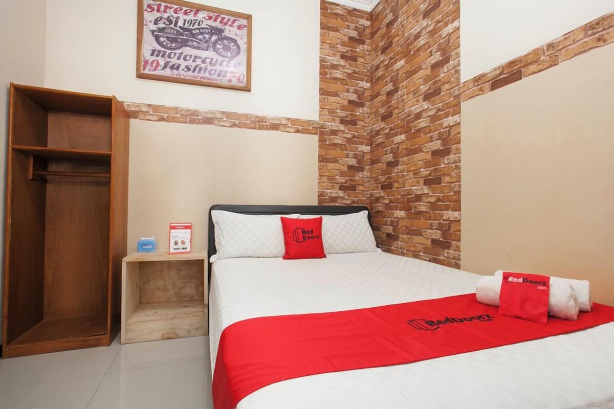 Une chambre d’hôtel avec un mur d’accent en briques, un lit avec une couverture et des oreillers rouges, une armoire en bois et une petite table de chevet avec une boîte et un oreiller.