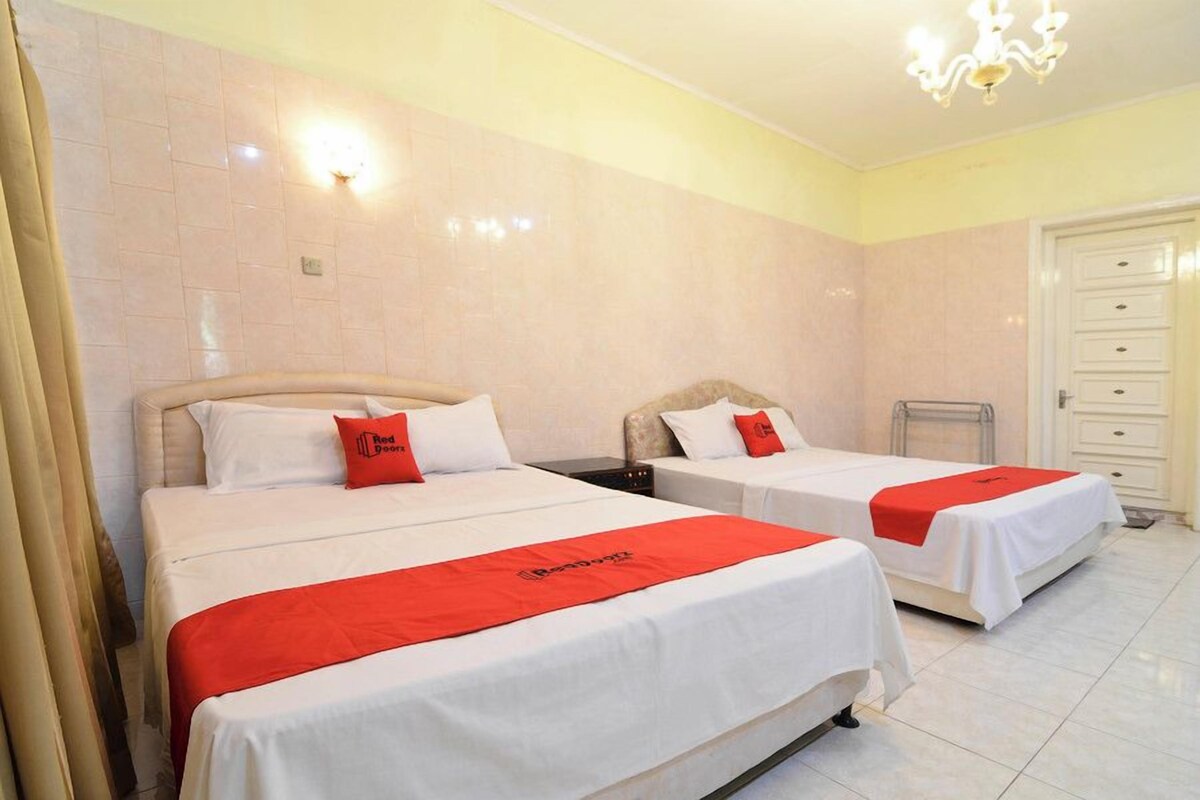 A hotel room with two beds, each with white bedding and red accents, a chandelier, and a tiled floor.