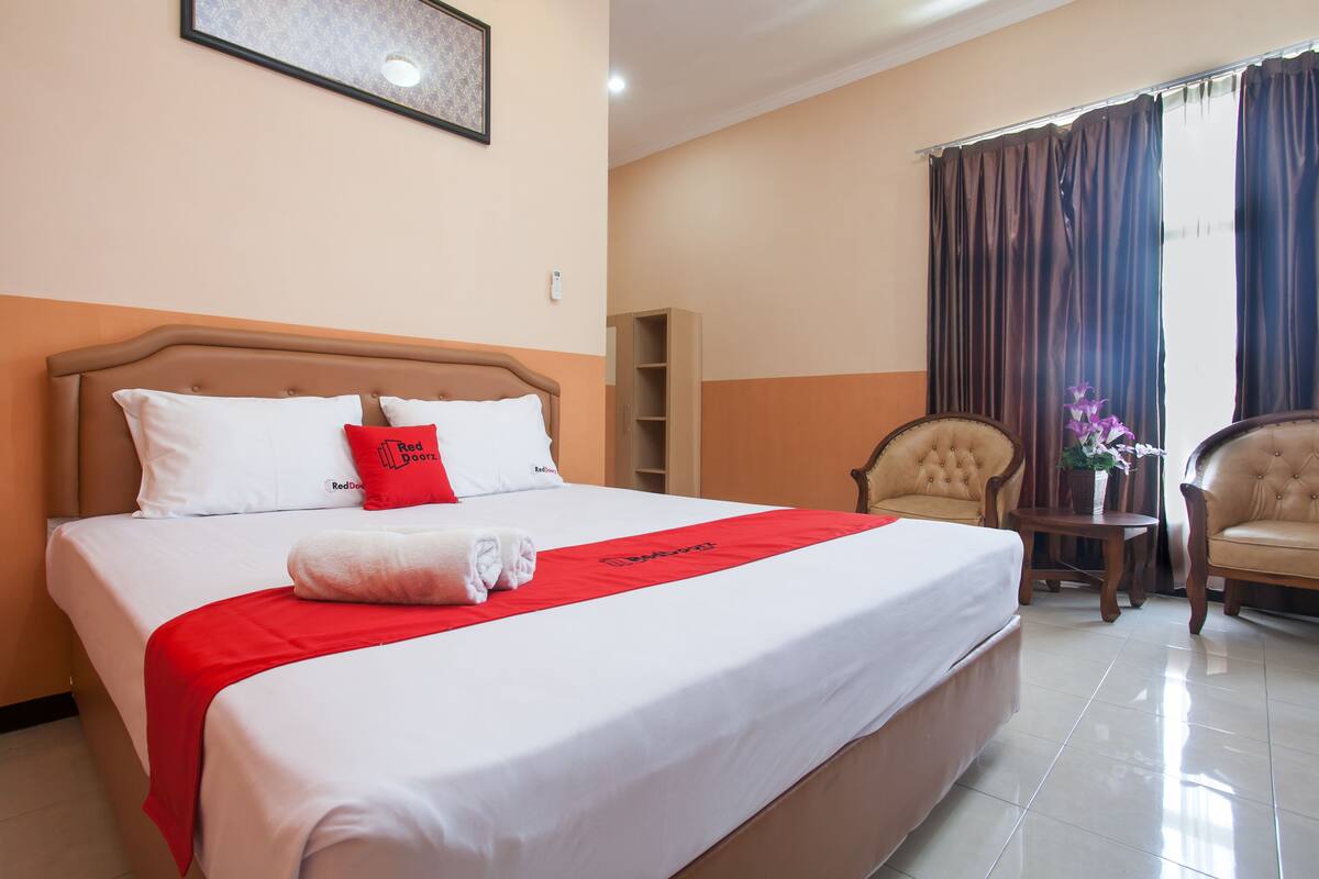 Quarto de hotel com uma cama grande, um cobertor vermelho e um travesseiro vermelho. Há uma pequena mesa com um vaso de flores e duas cadeiras.