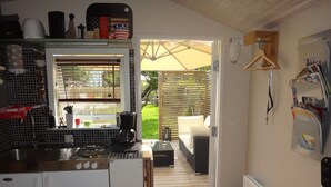 A compact kitchen with a sink, stove, and coffee maker. A small refrigerator is visible, and a window with blinds is present. The room leads to a patio with a sofa and a wooden deck. - Eklanda Studio 51 (Gothenburg)