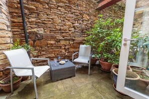 Un patio con paredes de piedra, dos sillas de mimbre, una mesita y varias macetas con plantas.