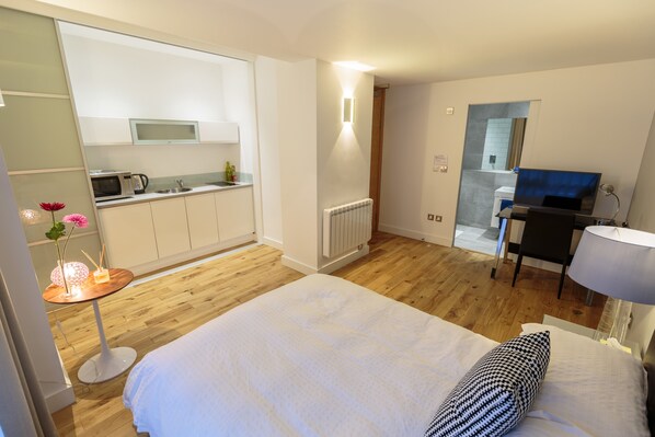 Habitación de hotel con cama, una mesita con flores, kitchenette, televisión y baño a la vista a través de una puerta abierta.