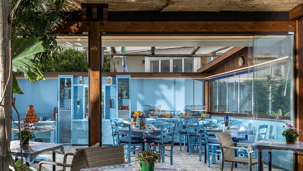 Un espace extérieur aménagé pour les repas, avec des chaises et des tables bleues, une pergola en bois et une vue sur la rue grâce à de grandes portes vitrées.