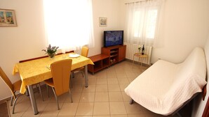 Una habitación pequeña y luminosa con una mesa de comedor, sillas, un televisor, un sofá y una pequeña mesa con una planta.